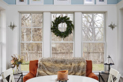 Sunroom remodeling project. Four season covered porch remodel. Light blue sunroom with winter scene in background. A white table with two chairs sits in the foreground. A star pendant chandelier hangs in the middle of the photo. A red couch is in the background with a throw blanket. 
