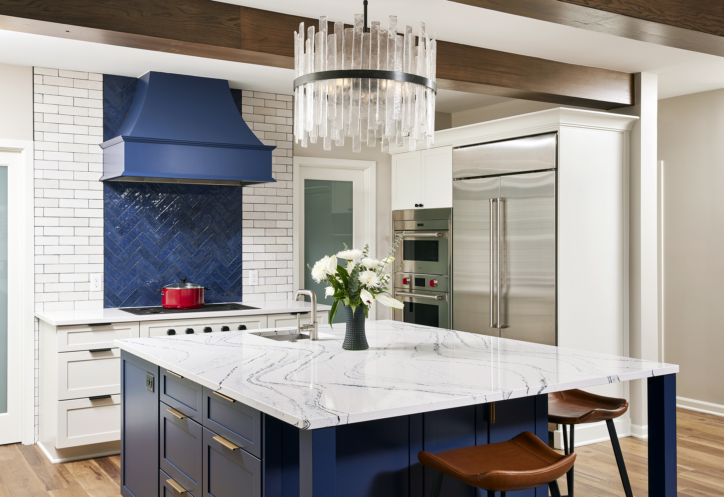 kitchen remodel with large, white square island, rich blue cabinetry, glazed blue backsplash tile and range hood, two ovens, fridge and island seating